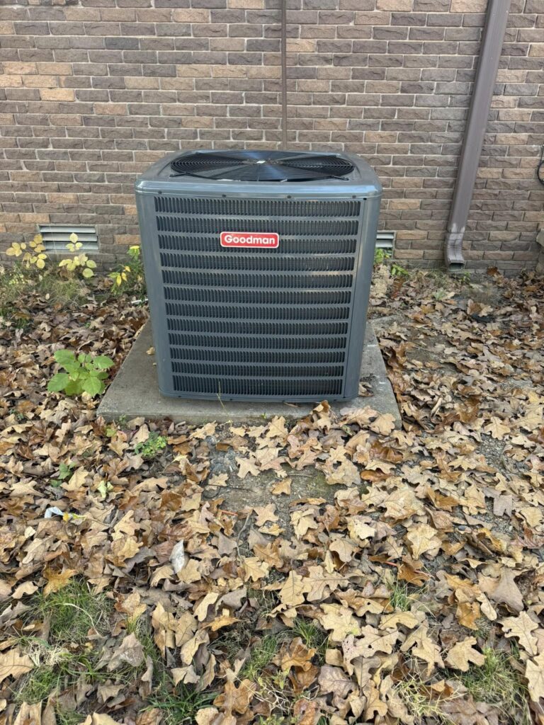 Goodman outdoor AC condenser unit on a concrete pad against a brick wall, surrounded by fallen leaves