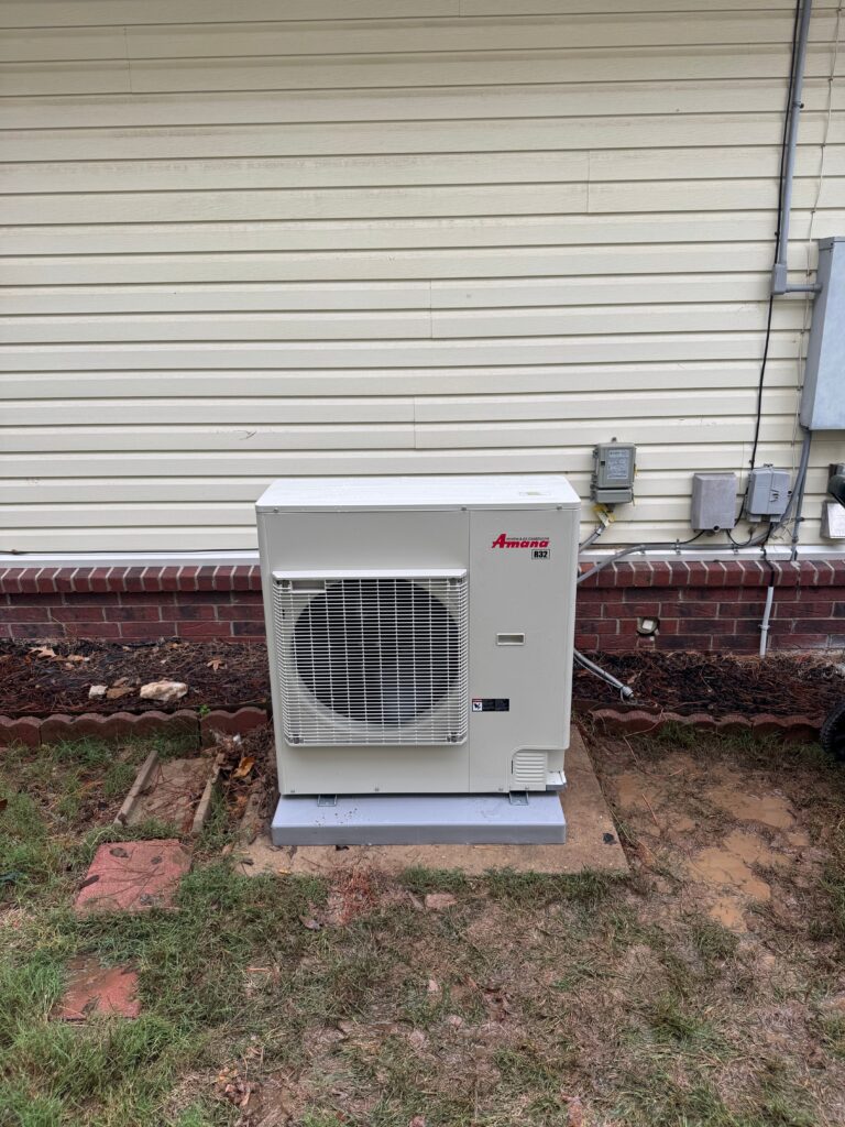 Amana R32 outdoor heat pump unit installed on a concrete pad beside a home's exterior wall