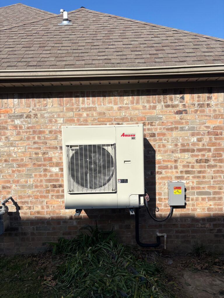 Amana R32 outdoor heat pump unit mounted on the exterior brick wall of a residential home