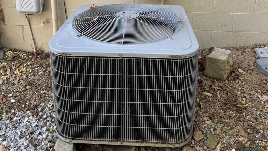 Older outdoor AC condenser unit with dirty coils and debris, placed on gravel beside a brick wall